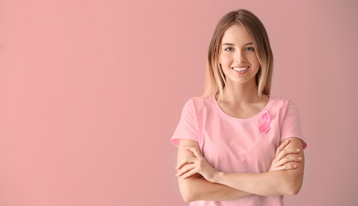 Smiling woman wearing a pink shirt with a ribbon on it, standing against a pink background.
