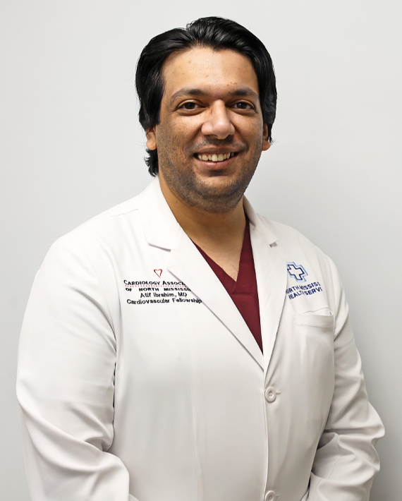 A smiling medical professional wearing a white coat with medical insignias standing against a plain background.