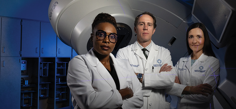 Three doctors stand in a radiation treatment room, highlighting a clinical oncology care team and equipment.