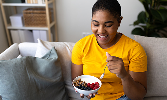 Person smiling while holding and eating from a bowl of fruits and granola on a couch.