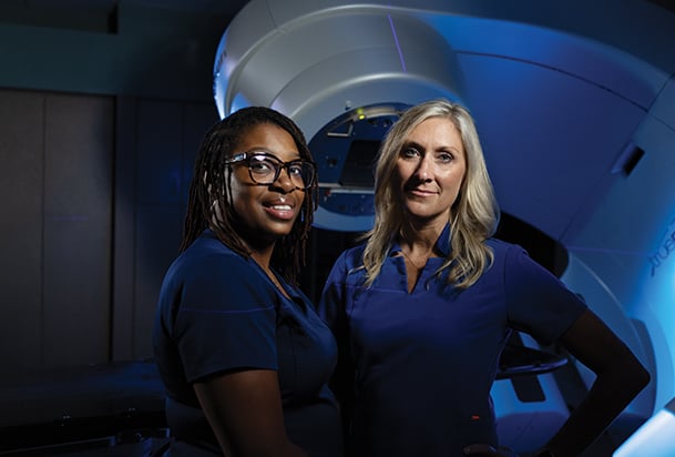 Two healthcare workers stand in front of a large radiation therapy machine in a dimly lit treatment room.