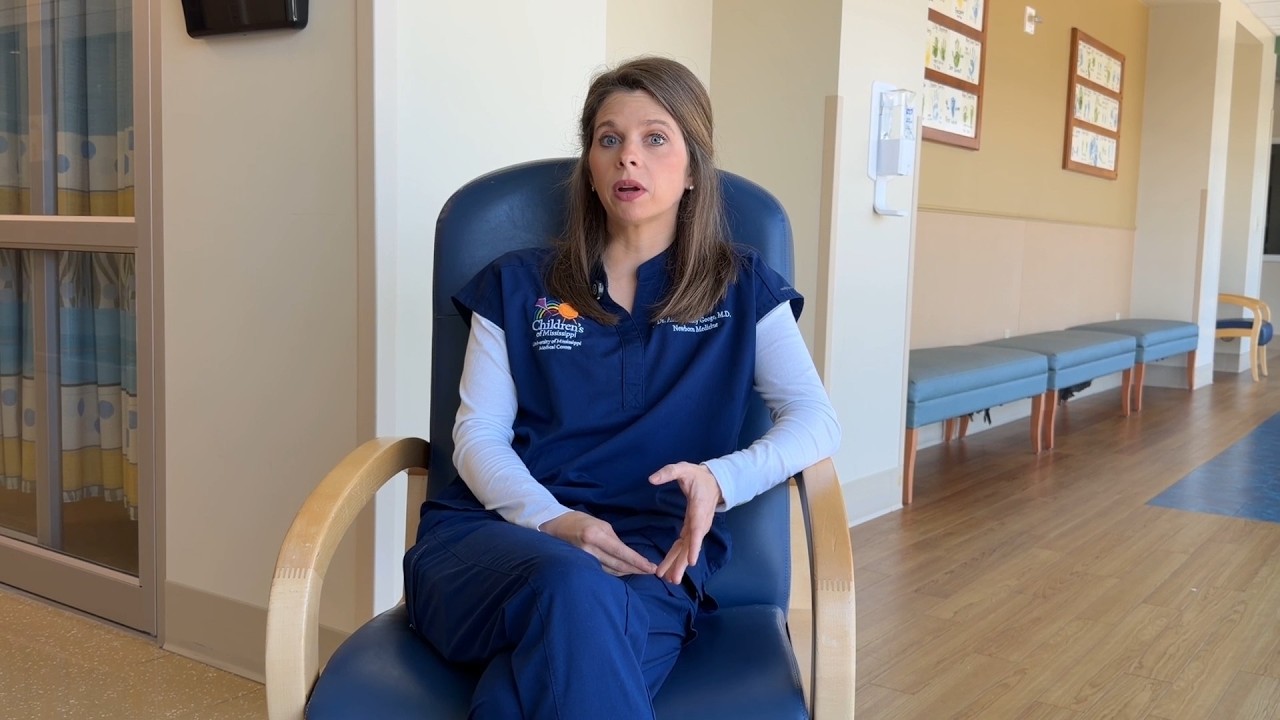 A woman in blue medical attire sits on a chair in a hospital setting, gesturing as if speaking.