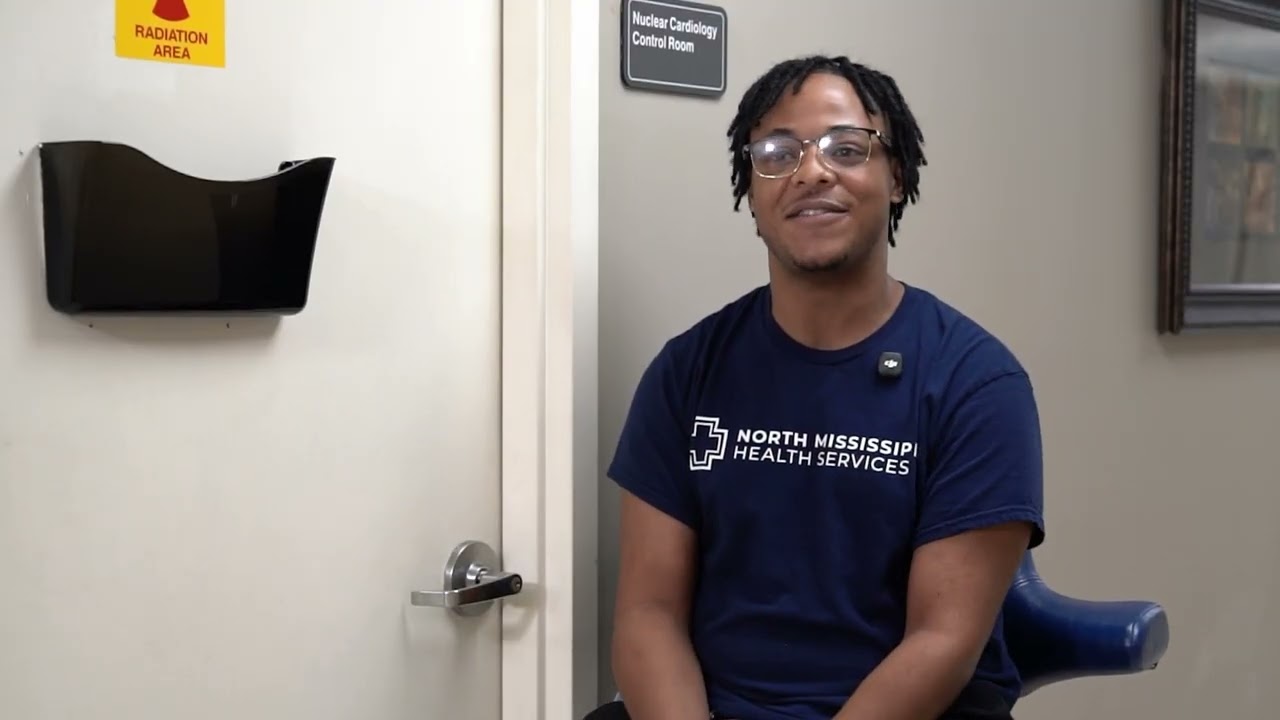 A smiling individual wearing a North Mississippi Health Services shirt seated next to a closed door with signage indicating a radiation area and a nuclear cardiology control room.