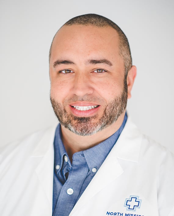 A healthcare professional smiling while wearing a white coat and a blue collared shirt.