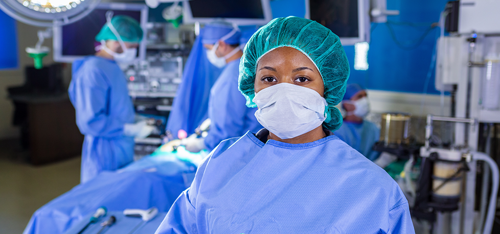 A focused medical professional wearing scrubs and a mask in an operating room with team members in the background.