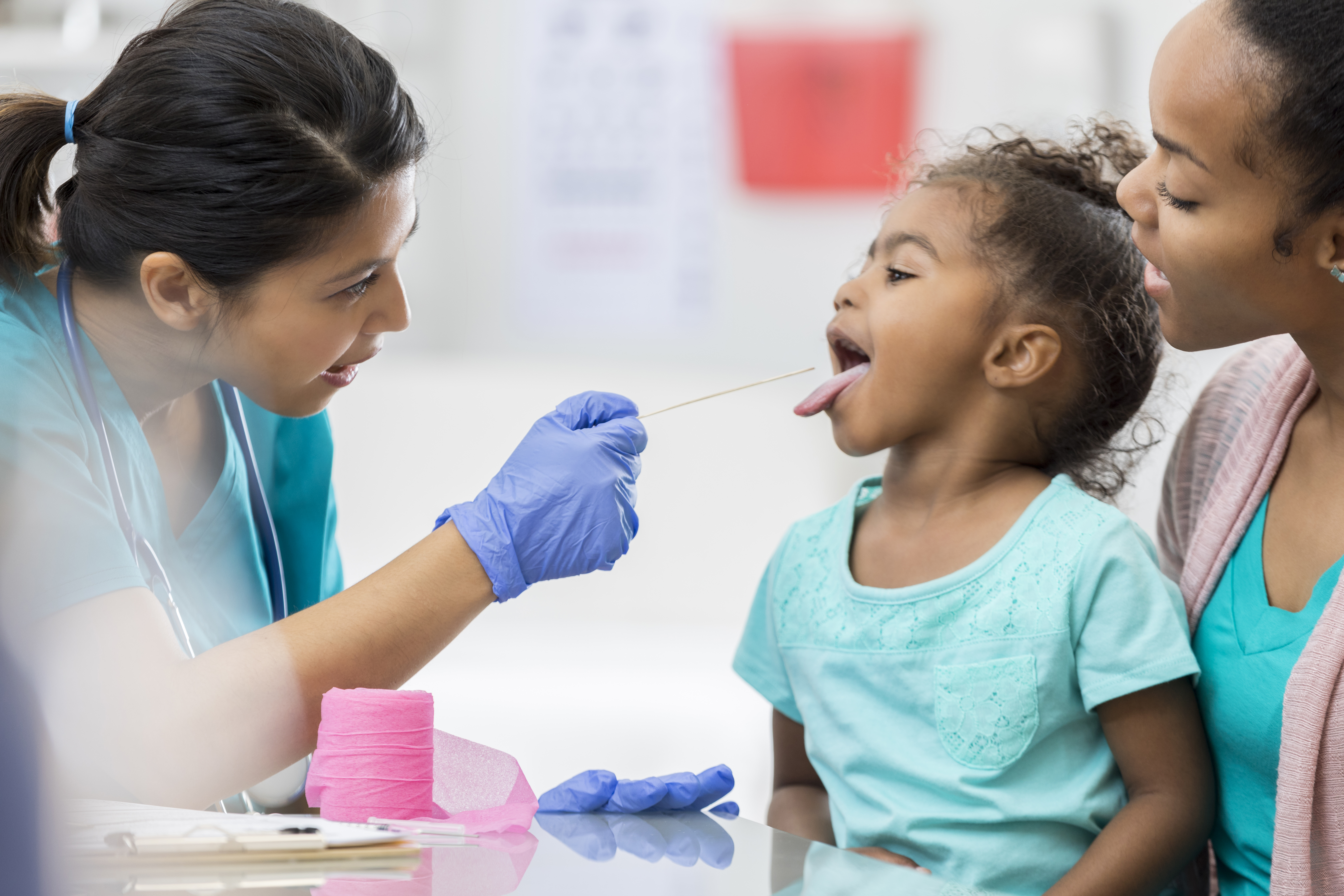 A healthcare professional wearing gloves uses a tongue depressor to examine a young girl's throat while an adult looks on.