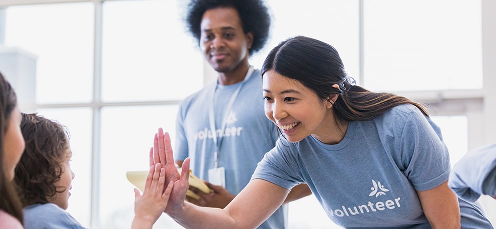 A smiling volunteer high-fives a child while another volunteer looks on in a bright indoor setting.