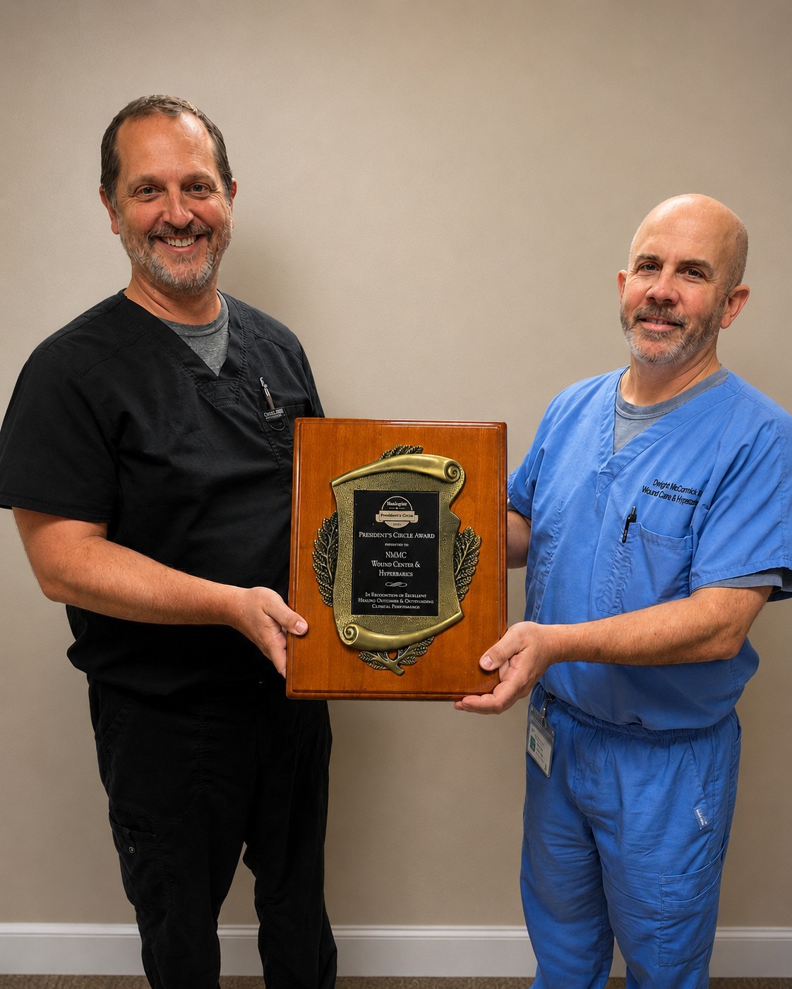 Two men in medical scrubs proudly holding a wooden award plaque and smiling.