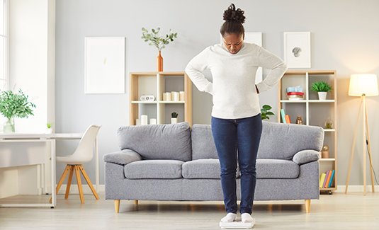 A person standing on a scale in a modern living room with a sofa and shelves in the background.