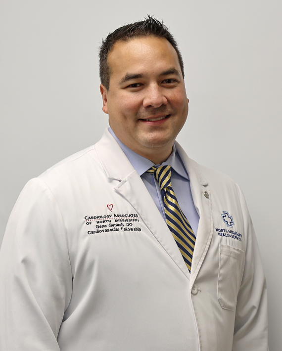 A medical professional wearing a white coat with embroidered text and a striped tie, smiling in front of a neutral background.
