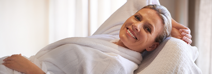 Smiling woman relaxing in a white robe while lying on a cushioned surface.