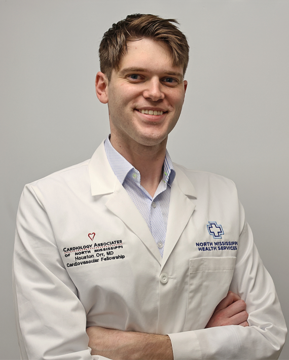 A person smiling, wearing a white medical coat with institutional logos.